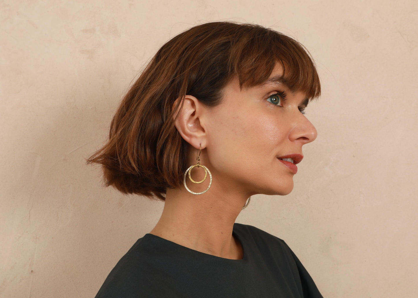 A woman with a short brown bob hairstyle is shown in profile, wearing a pair of gold double-ring hoop earrings against a textured beige wall.