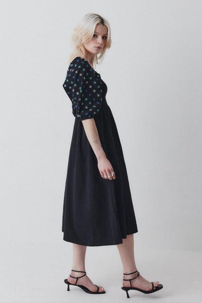 A woman modeling a black dress with hand-embroidered details on the sleeves, a flattering shirred bodice, and side pockets.