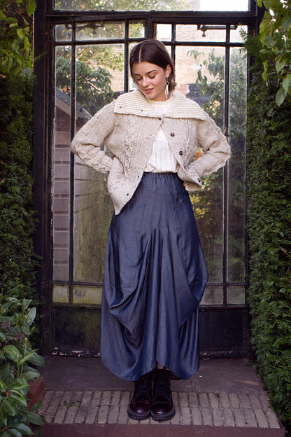 Woman wearing a textured sweater and blue skirt standing in front of a glass door with greenery around.