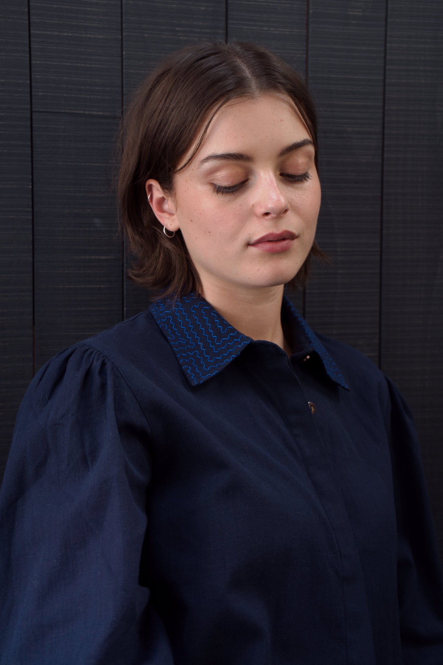 Woman wearing a dark blue shirt with a textured collar against a black wooden panel background