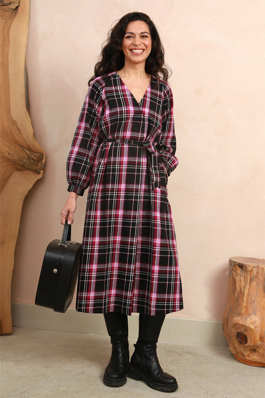 A woman stands indoors wearing a long-sleeve plaid wrap dress with a black, pink, and white tartan pattern, paired with black boots and holding a black suitcase.