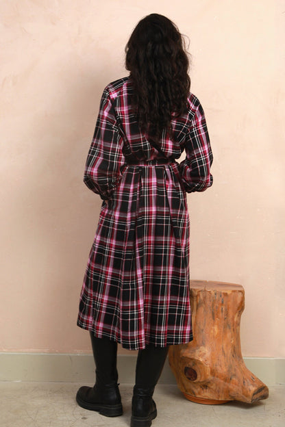 A woman viewed from behind wearing a long-sleeved plaid wrap dress in black, pink, and white, standing next to a rustic wooden stool against a textured beige wall.