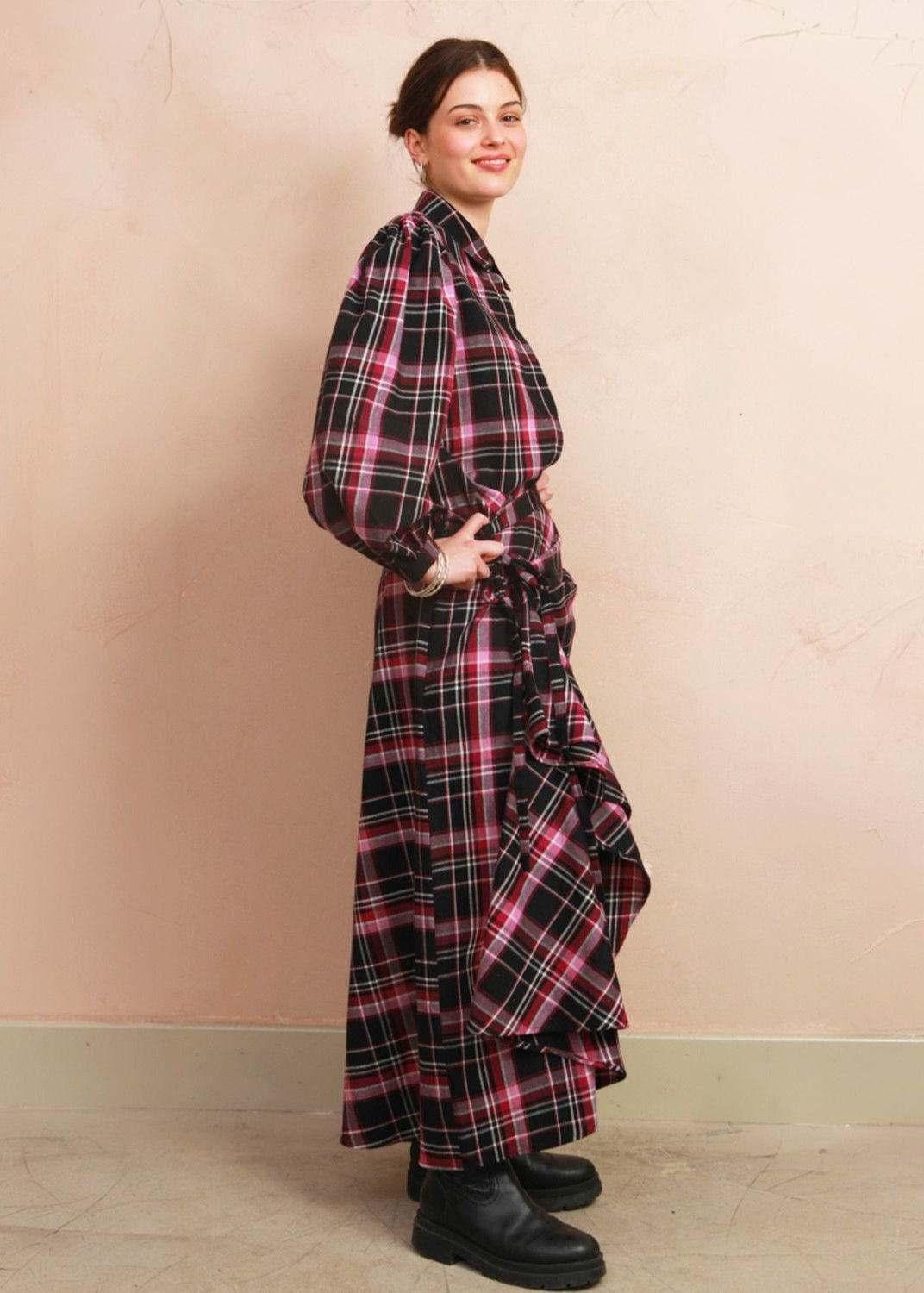 A woman modeling a long-sleeved, collared plaid wrap dress with a ruffled hem, standing against a textured peach-pink wall.
