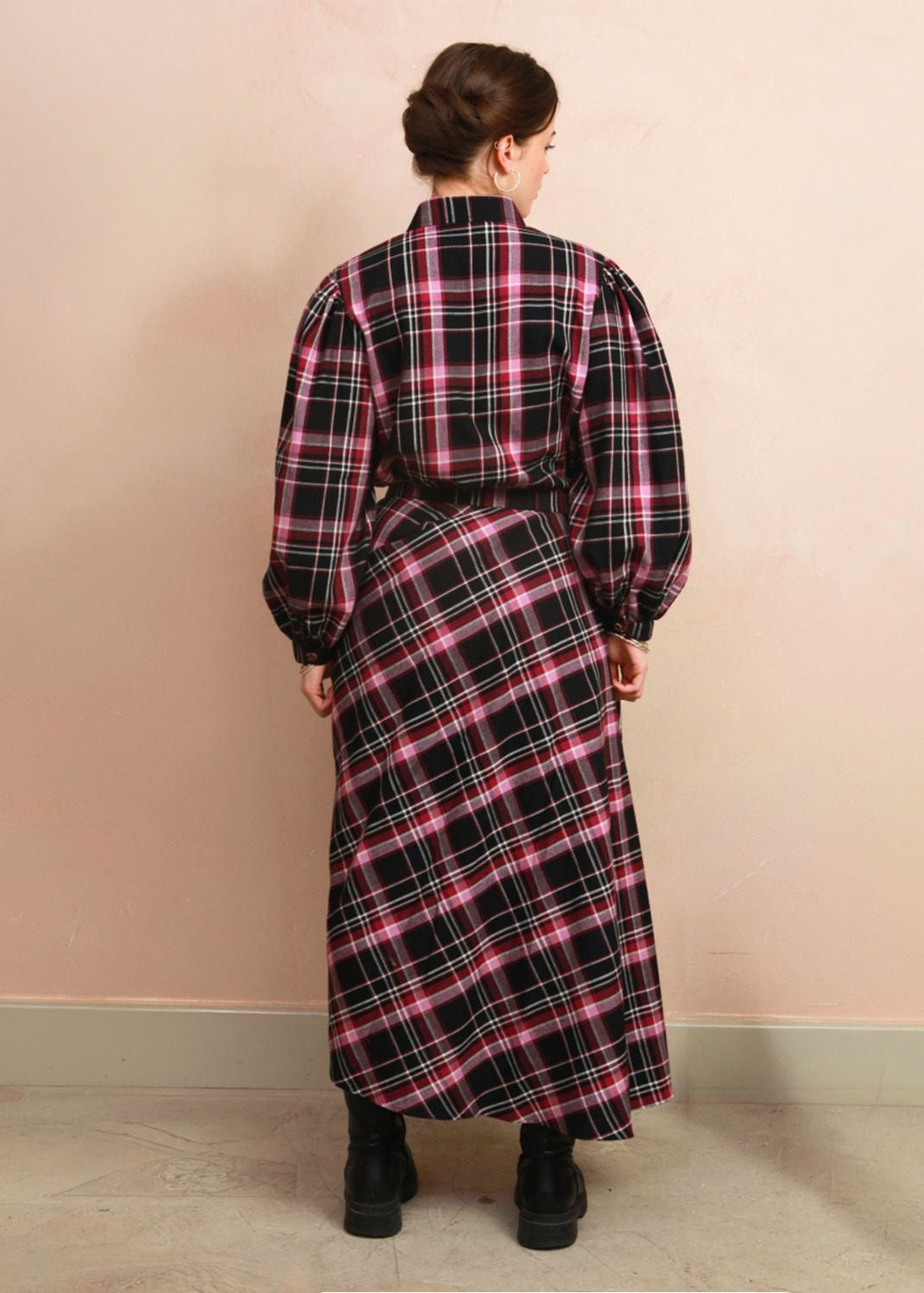 A woman viewed from behind wearing a long-sleeved plaid shirt dress in black, pink, and white, standing against a pale pink wall.