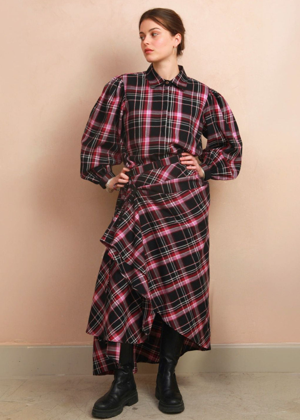 A model stands against a beige wall wearing a long-sleeved, high-waisted plaid shirt dress with a ruffled asymmetrical hem. The dress features a black base with pink and white plaid stripes, styled with black boots.