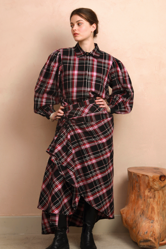 A model stands against a beige wall wearing a long-sleeved, high-waisted plaid skirt with a ruffled asymmetrical hem. This features a black base with pink and white plaid stripes, styled with black boots.