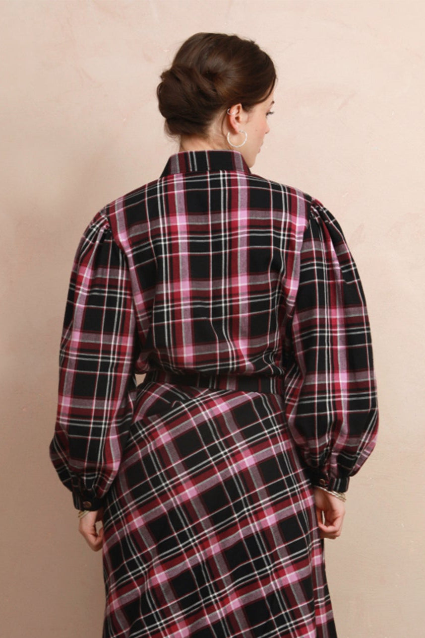 A woman viewed from behind wearing a long-sleeved black plaid shirt with pink and white stripes, standing in front of a beige wall.