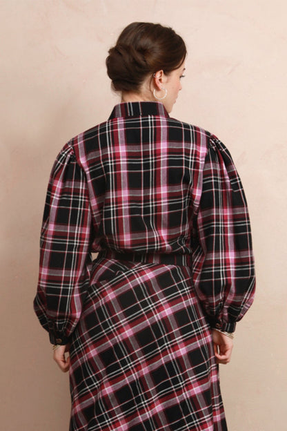 A woman viewed from behind wearing a long-sleeved black plaid shirt with pink and white stripes, standing in front of a beige wall.