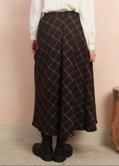 A person shown from the back wearing a black skirt with a white windowpane plaid pattern, paired with a white blouse and black boots, standing against a pale pink wall.