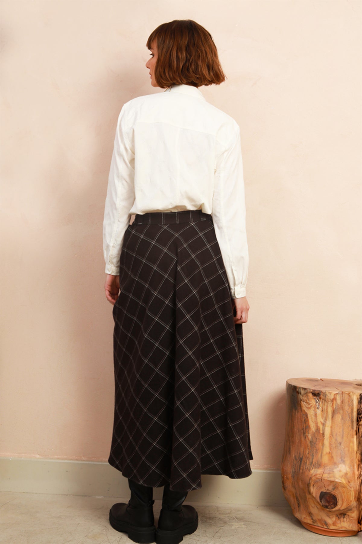 A woman is shown from behind wearing a white long-sleeve button-up shirt and a black high-waisted pleated skirt with a subtle diagonal grid pattern. She is standing in front of a light beige wall next to a rustic wooden stool.