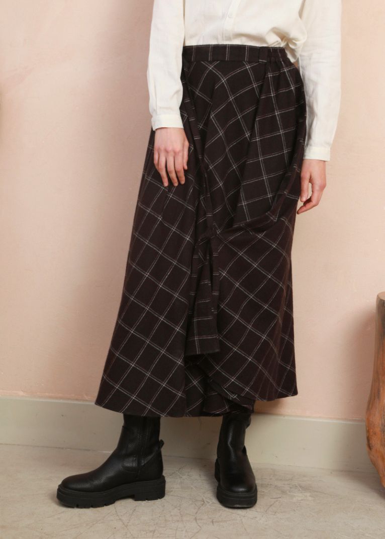A person wearing a dark black plaid wrap midi skirt with a white long-sleeve top and black boots, standing against a textured beige wall.