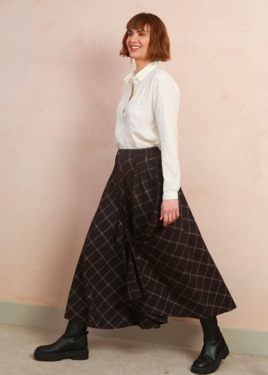 A woman wearing a white long-sleeve button-up shirt and a black plaid skirt, paired with black leather boots, standing against a textured light pink wall.
