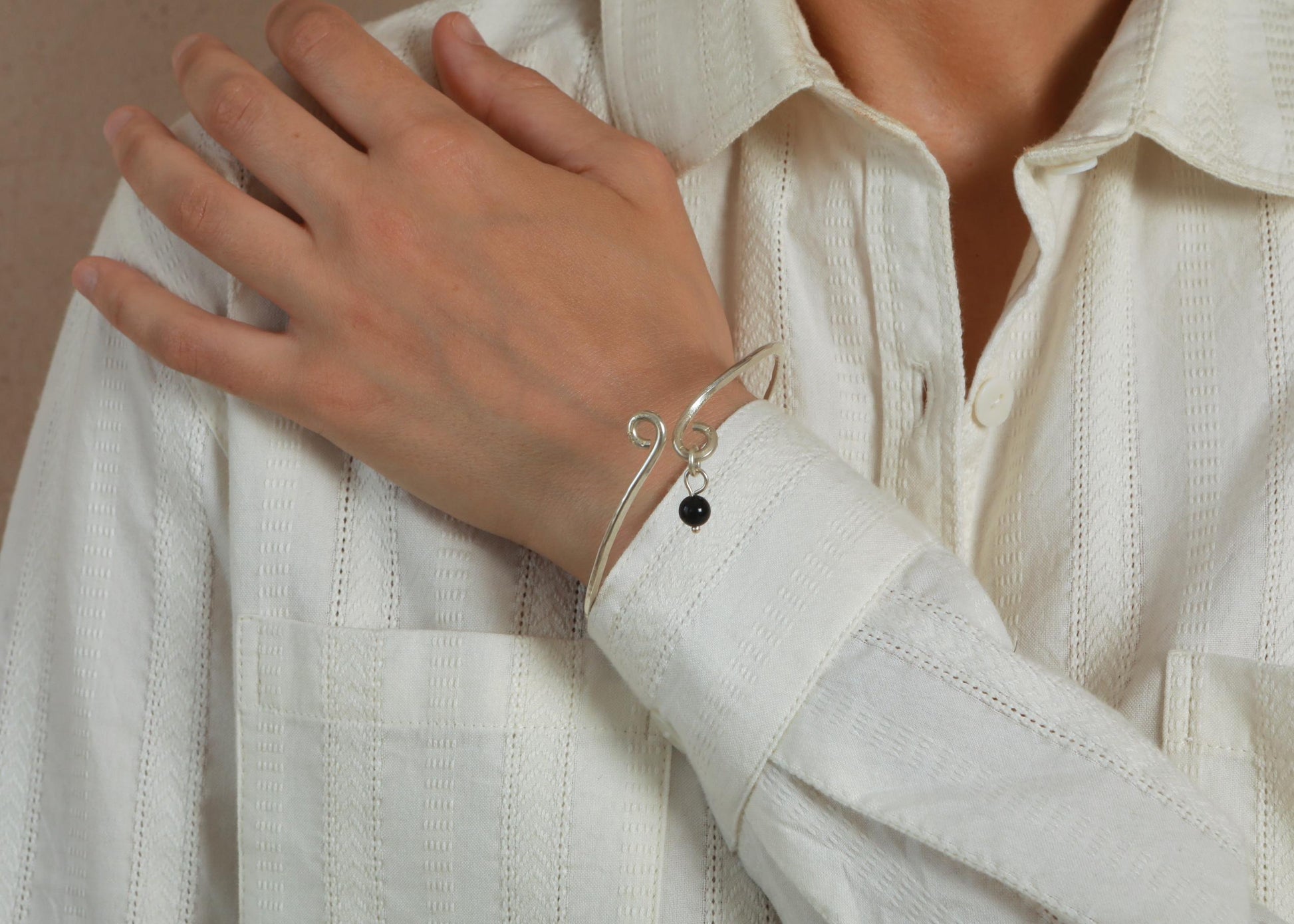 A close-up of a person wearing a delicate silver wire bangle bracelet with a small black bead, paired with a white textured button-up shirt.