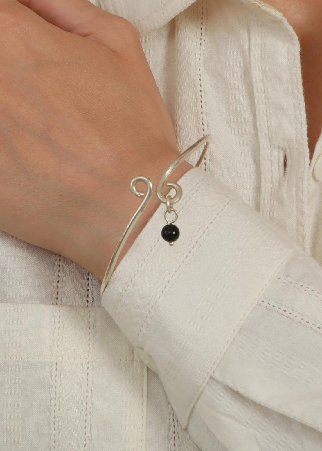 A close-up of a person wearing a delicate silver charm bracelet with a small black bead, paired with a white textured button-up shirt.