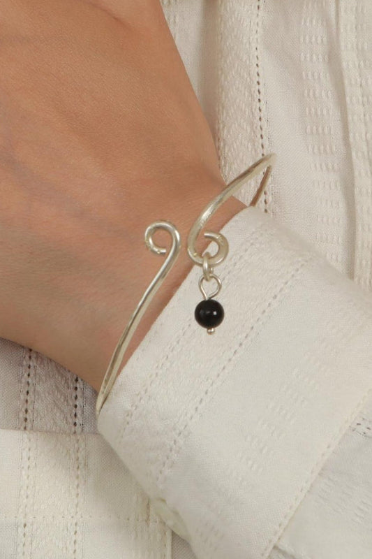 A close-up of a person wearing a delicate silver charm bracelet with a small black bead, paired with a white textured button-up shirt.