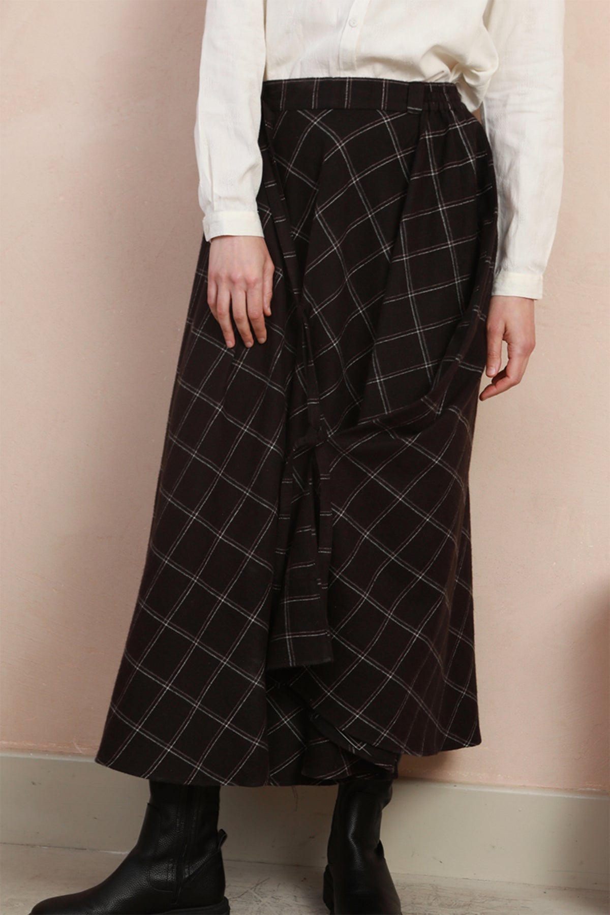 A person wearing a dark black plaid wrap skirt with a white long-sleeve top and black ankle boots, standing against a pale pink wall.