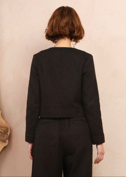 A woman viewed from behind wearing a black textured cropped jacket and matching wide-leg trousers against a warm beige wall.