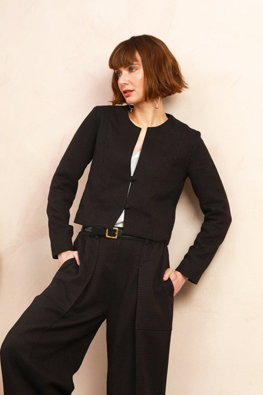 A woman modeling a black cropped jacket and wide-leg trousers set with a white top and black shoes, posing against a light beige wall next to a large wooden slab.