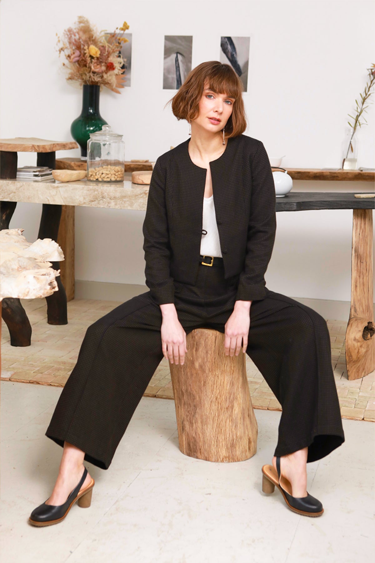 A woman sits on a rustic wooden stump in a styled indoor setting, wearing a black tailored jacket and matching wide-leg trousers over a white top, with black mule heels.