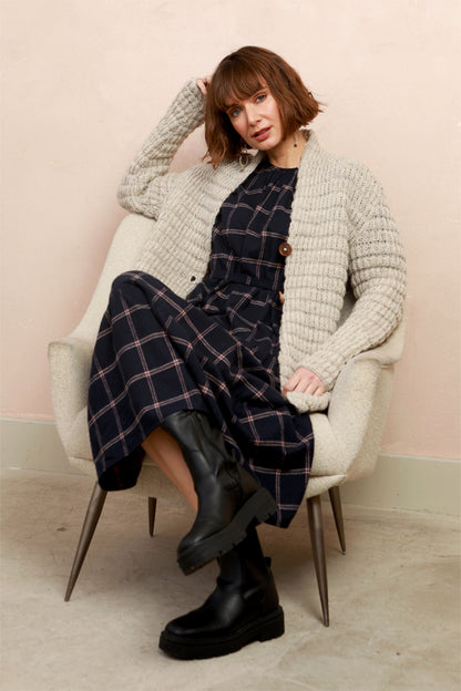 A woman wearing a navy blue plaid midi dress with long sleeves and a tiered skirt, sat down on chair