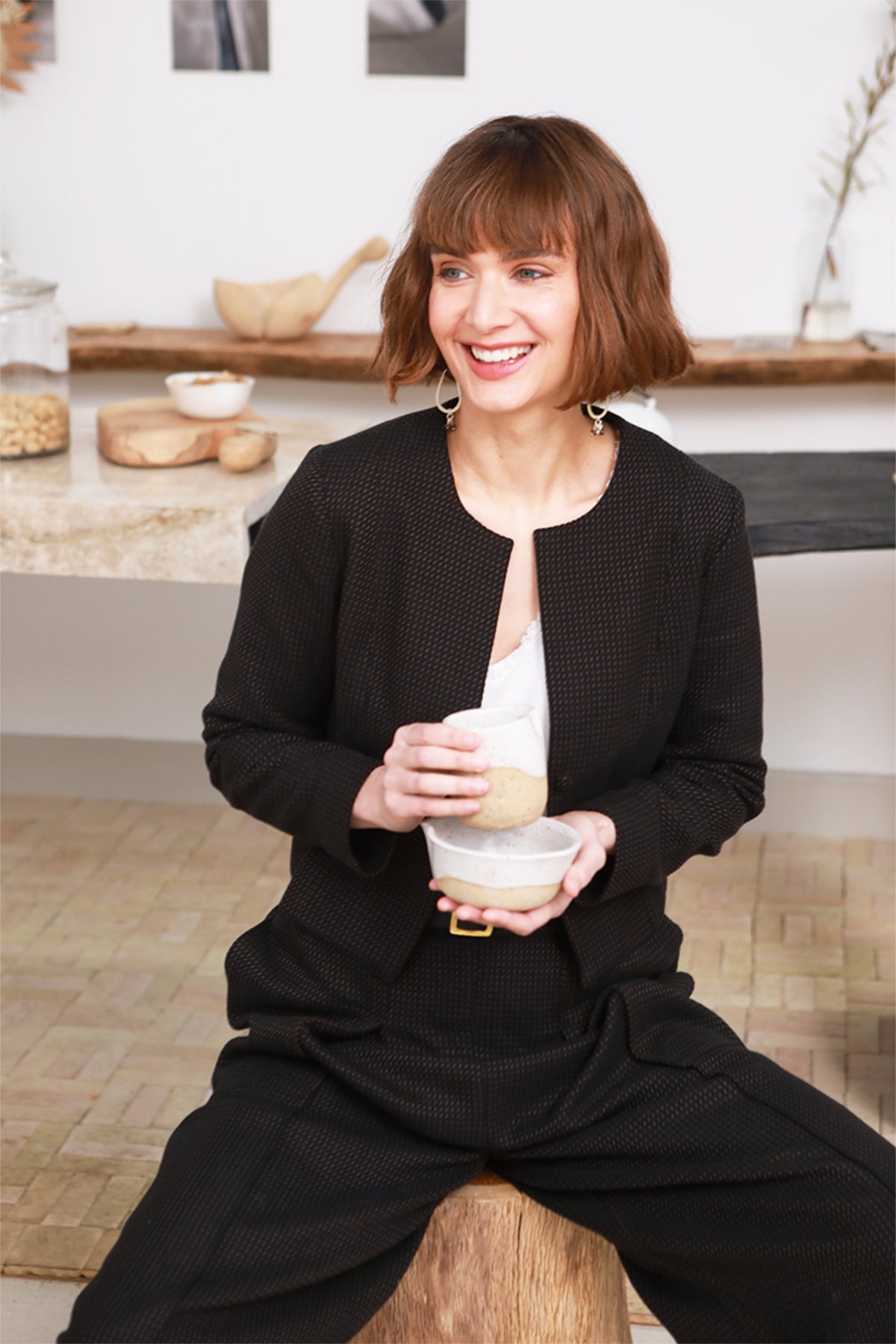 A woman with short brown hair sits smiling on a wooden stool in a bright kitchen, wearing a black textured jacket and matching wide-leg pants over a white top, while holding a ceramic mug.