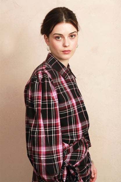 A woman wearing a black, pink, and white plaid button-down shirt, photographed from the side against a textured beige background.
