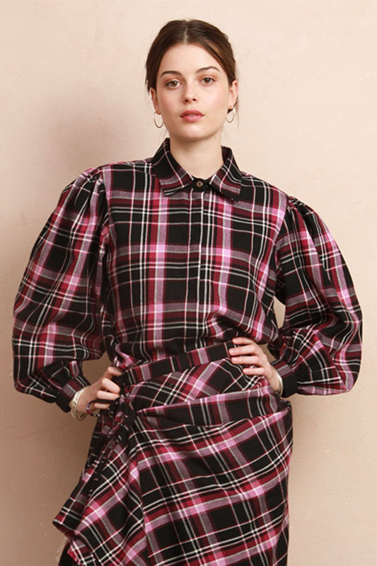 A model stands in a studio wearing a long-sleeved black and pink plaid shirt and skirt with an asymmetrical ruffled hem, paired with black boots. She is posed next to a rustic wooden stool against a beige wall.
