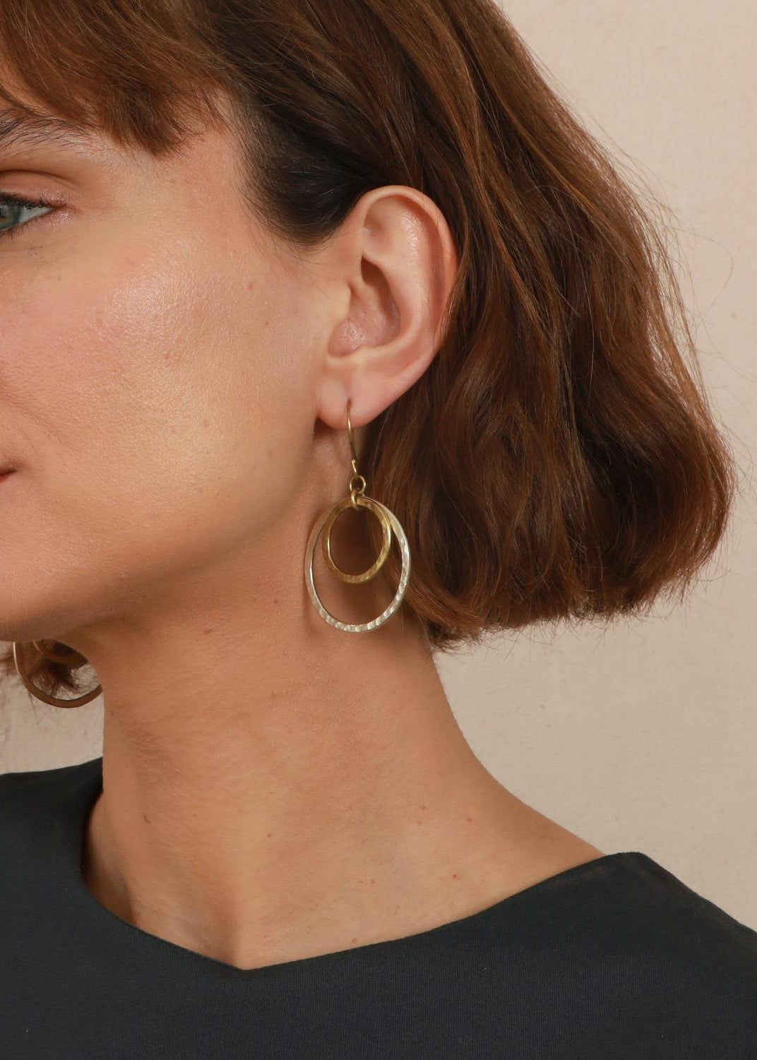 A woman with a short brown bob haircut and bangs is shown in profile, wearing large gold and silver hoop earrings and a dark charcoal top against a textured beige wall.
