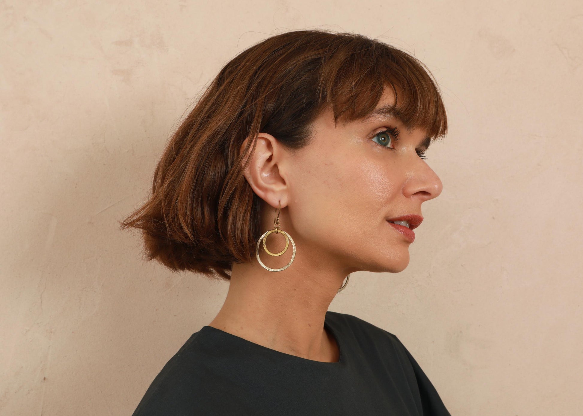 A woman with a short brown bob hairstyle is shown in profile, wearing a pair of gold double-ring hoop earrings against a textured beige wall.