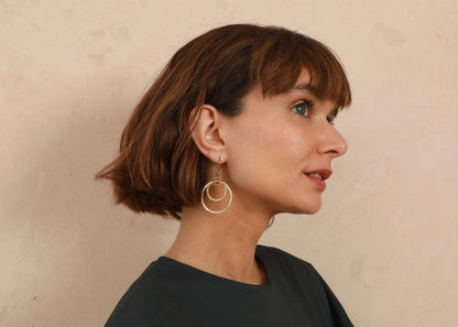 A woman with a short brown bob hairstyle is shown in profile, wearing a pair of gold double-ring hoop earrings against a textured beige wall.