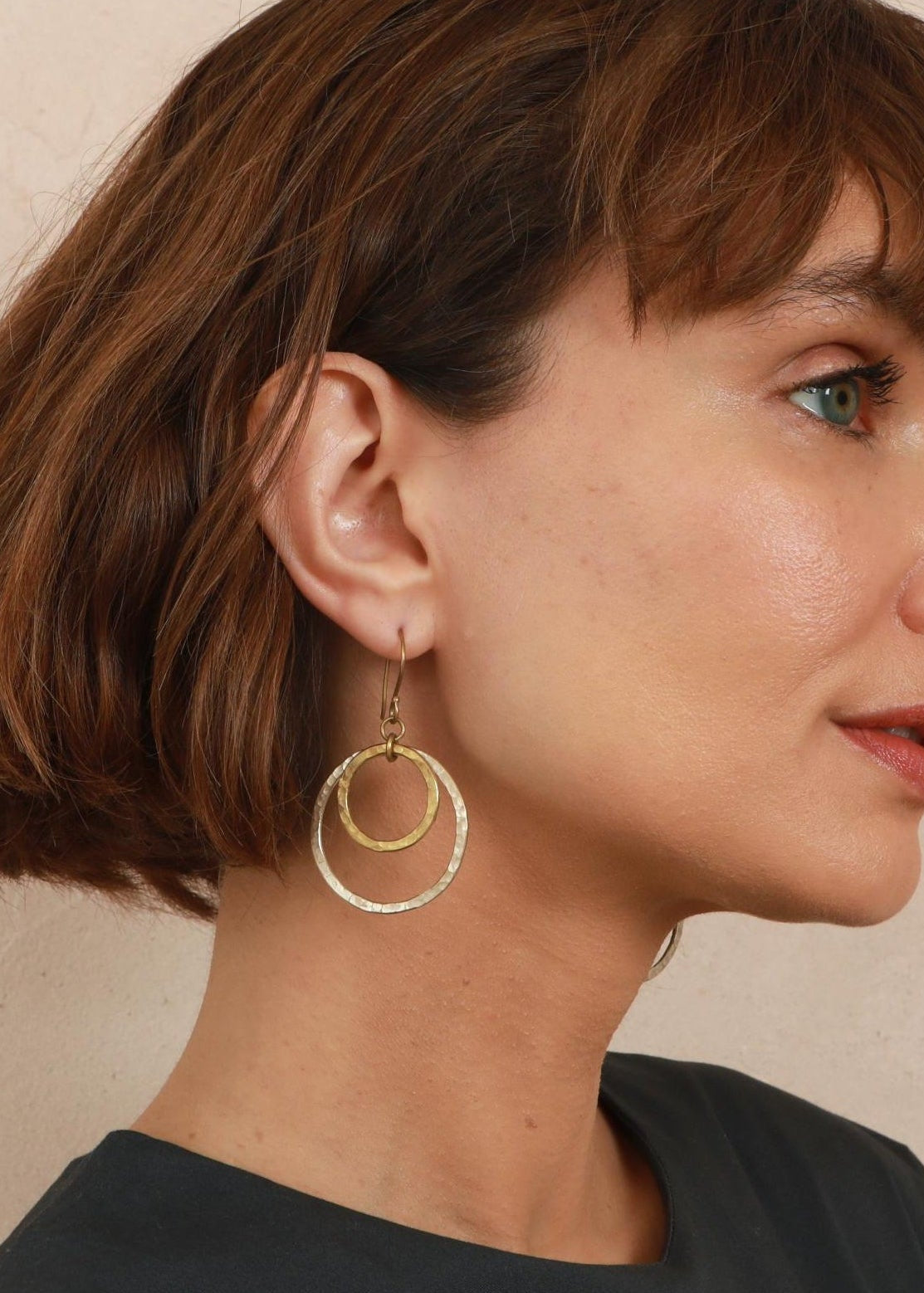 A woman with a brown bob haircut is shown in profile wearing a pair of gold double-ring hoop earrings against a neutral beige background.