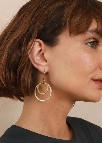 A woman with a brown bob haircut is shown in profile wearing a pair of gold double-ring hoop earrings against a neutral beige background.