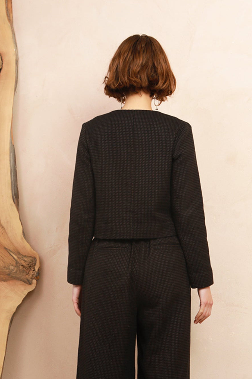 A model viewed from behind wearing a black long-sleeve cropped jacket and matching wide-leg trousers, standing against a light beige wall next to a large wood slab.