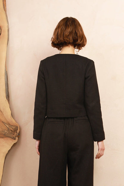 A model viewed from behind wearing a black long-sleeve cropped jacket and matching wide-leg trousers, standing against a light beige wall next to a large wood slab.