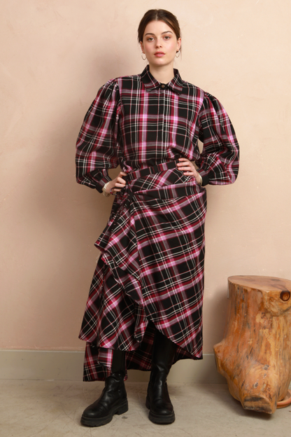 A model stands against a beige wall wearing a long-sleeved plaid shirt and skirt with a black, pink, and white tartan pattern, styled with black boots.