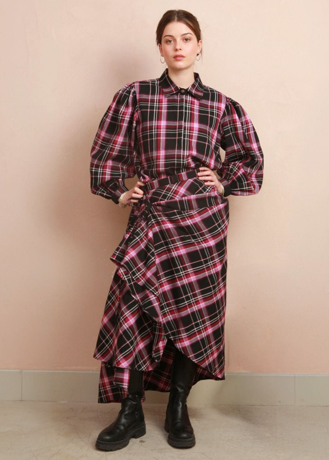 A model stands against a beige wall wearing a long-sleeved plaid shirt dress with a black, pink, and white tartan pattern, styled with black boots.