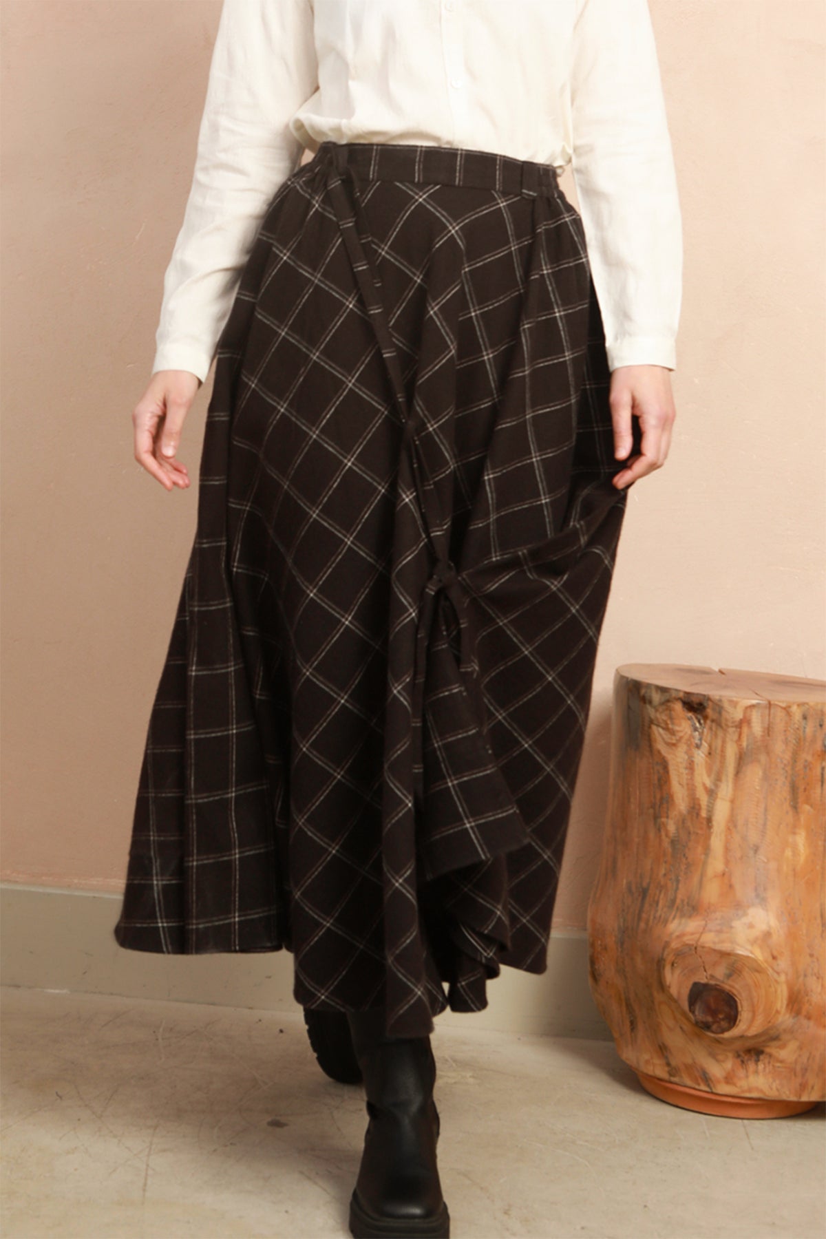 A person wearing a black and white plaid wrap skirt with a white blouse and black boots, standing next to a wooden stool against a beige wall.