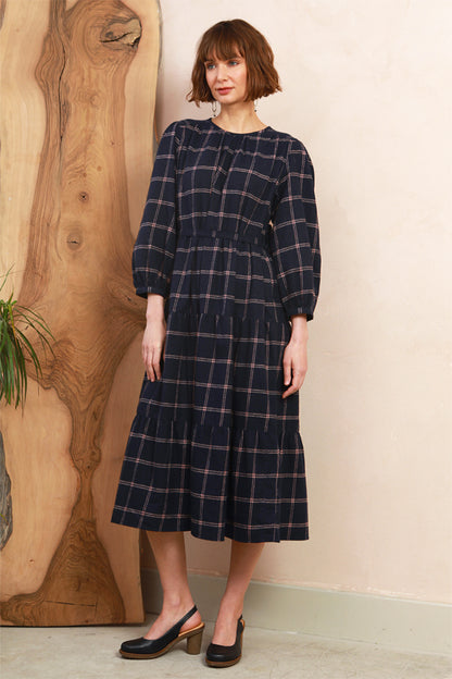 A woman wearing a dark navy blue tiered midi dress with a subtle pink plaid pattern, standing in front of a beige wall next to a large wooden slab.