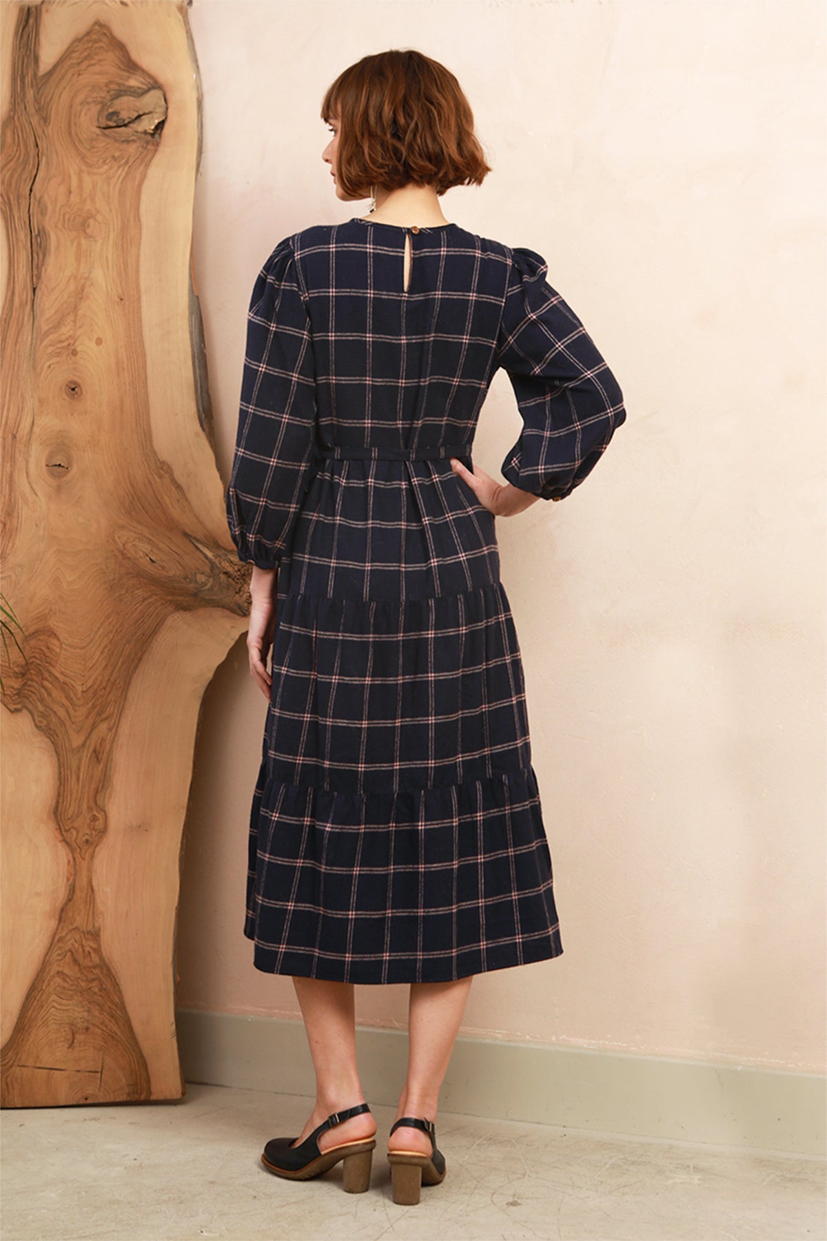 A woman wearing a navy blue plaid midi dress with long sleeves and a tiered skirt, shown from the back. She is standing in front of a beige wall next to a large wooden slab.