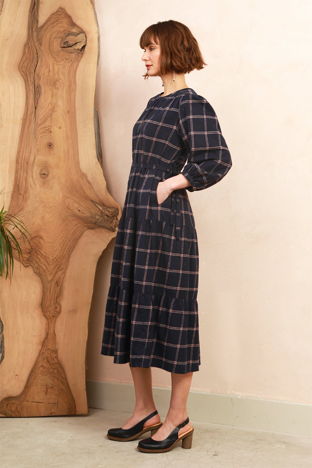 A woman wearing a navy blue plaid midi dress with three-quarter sleeves and side pockets, standing in a room with a large wooden slab decor piece and a beige wall.