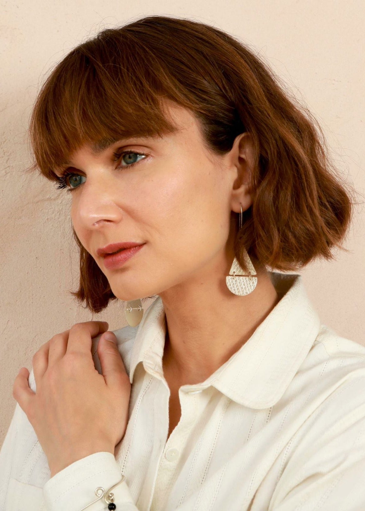 A woman with a short brown bob haircut and bangs, wearing a white collared shirt and a large textured silver dangle earring, posing against a light beige wall.
