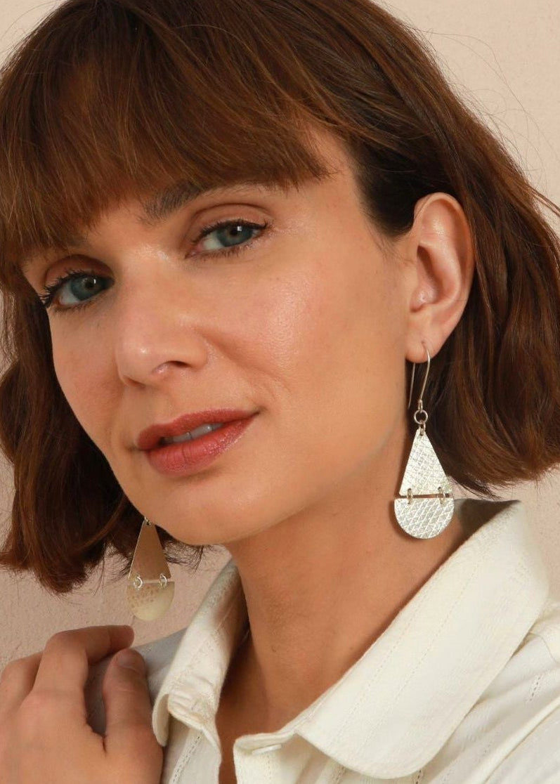 A close-up of a woman with brown hair wearing a silver teardrop-shaped textured dangle earring and a white collared shirt.