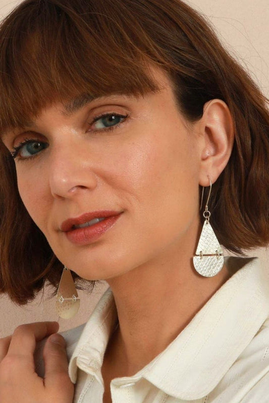 A close-up of a woman with brown hair wearing a silver teardrop-shaped textured dangle earring and a white collared shirt.
