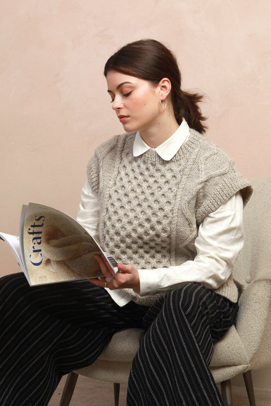 Seated model wearing the natural light grey hand-knit cable vest layered over a white shirt reading a magazine