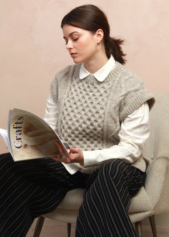 Seated model wearing the natural light grey hand-knit cable vest layered over a white shirt reading a magazine
