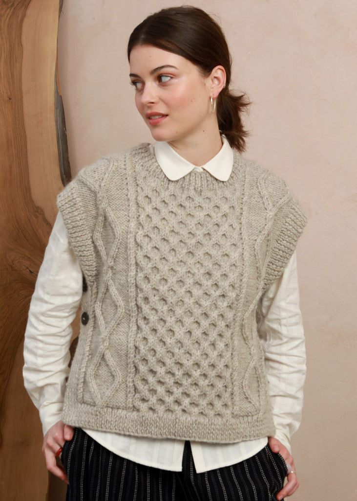 Front view of model wearing the natural light grey cable-knit slipover vest holding a book