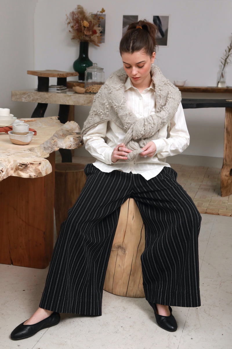 A woman sits on a wooden stool in a rustic indoor setting, wearing a white button-up shirt, a chunky light gray faux-fur collar, and wide-leg black pinstripe trousers with black flats.