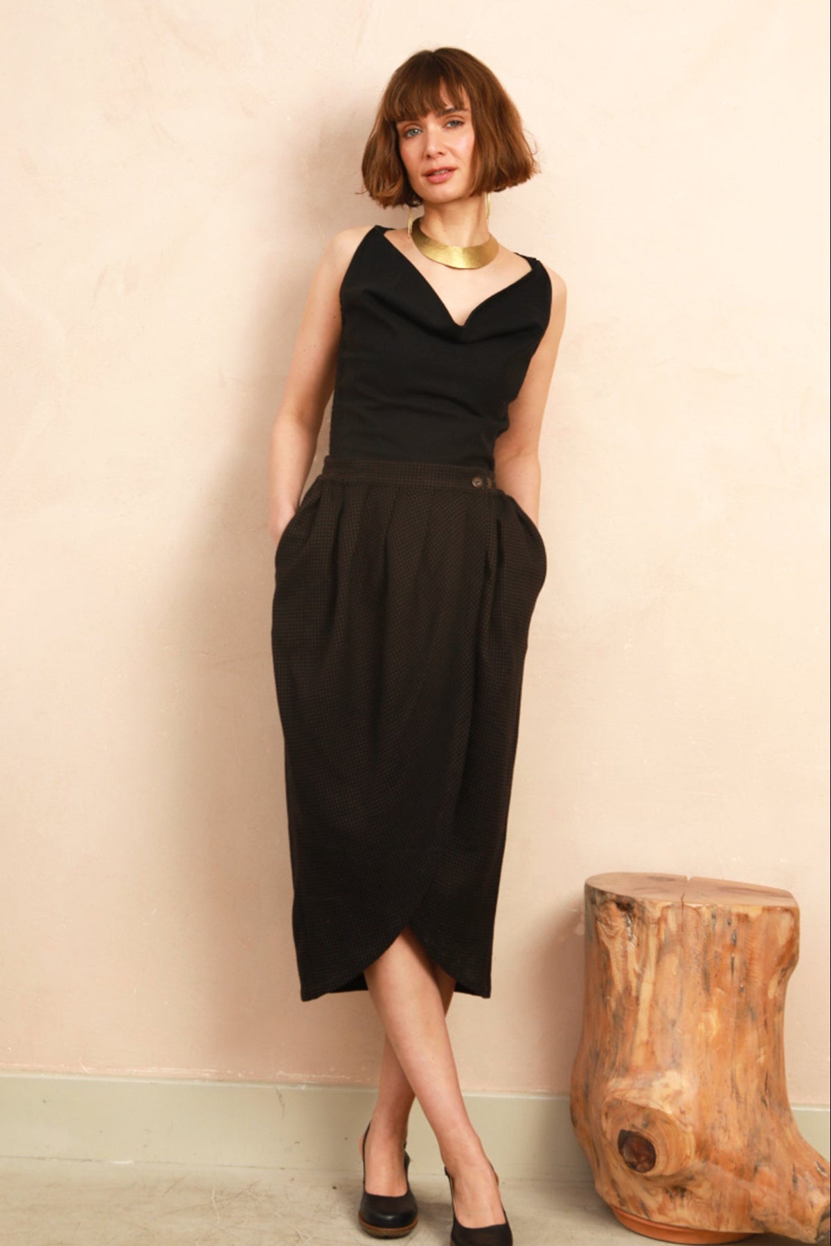 A woman with a short brown bob stands against a textured beige wall wearing a sleeveless black midi dress with a draped neckline, side pockets, and a slight slit. She is accessorized with a chunky gold necklace and black heels, with a rustic wooden stump stool to her right.