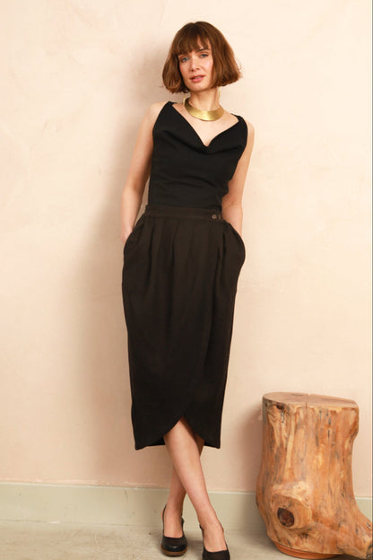 A woman with a short brown bob stands against a textured beige wall wearing a sleeveless black midi dress with a draped neckline, side pockets, and a slight slit. She is accessorized with a chunky gold necklace and black heels, with a rustic wooden stump stool to her right.