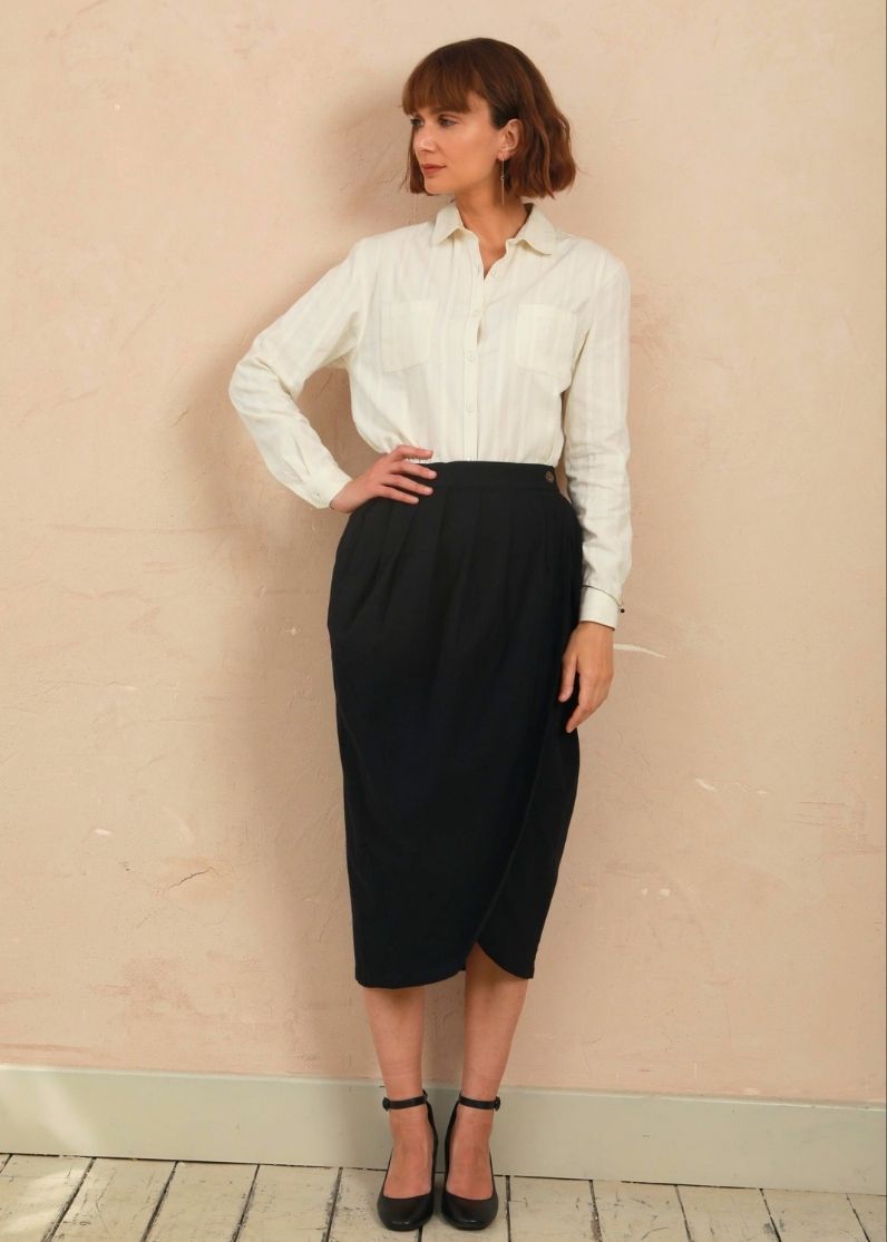A woman stands against a textured beige wall wearing a white long-sleeve button-up shirt tucked into a high-waisted black midi skirt, paired with black ankle-strap heels.